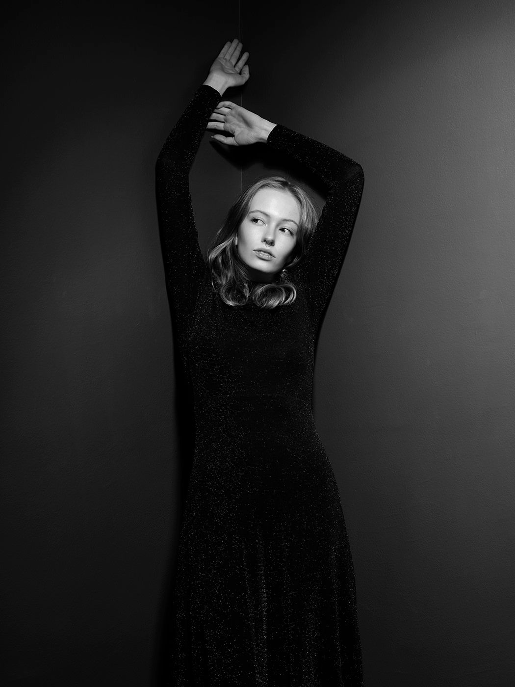 Black and white portrait of a young woman in a dark long-sleeve dress, arms raised gracefully overhead.