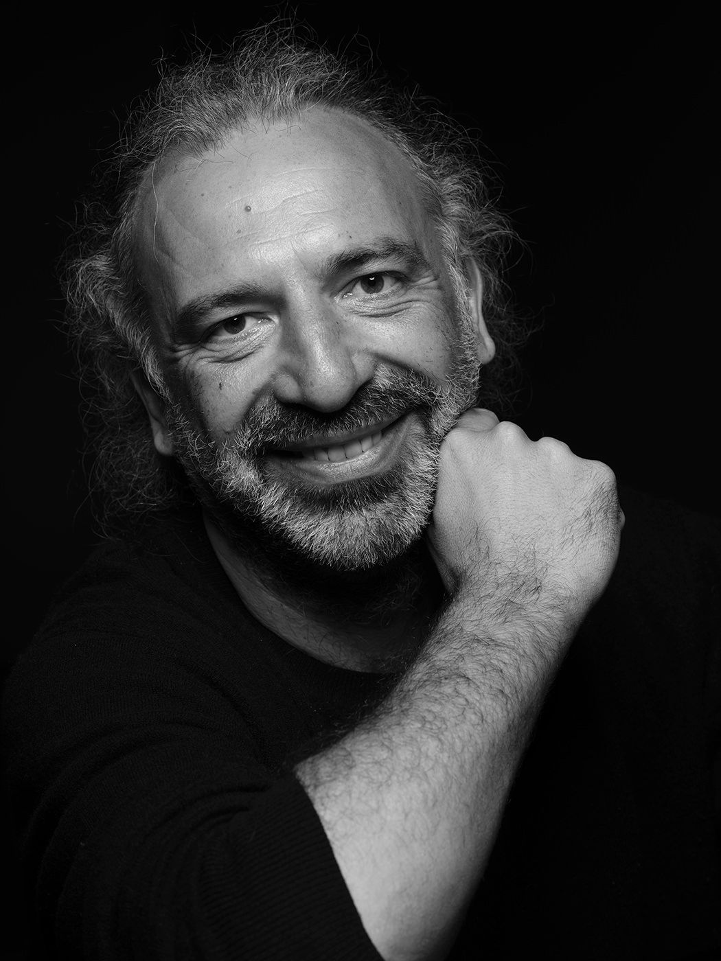 Black and white portrait of a smiling middle-aged man with curly hair and beard, resting chin on hand. Stefano Bolliani, pianist