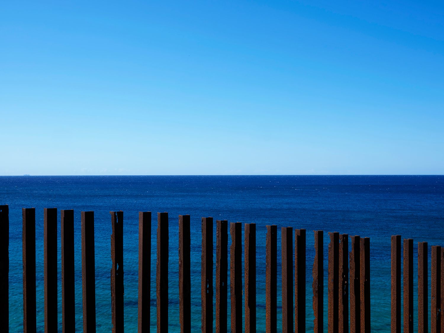 Wooden fence posts along a serene ocean shoreline under a clear blue sky.