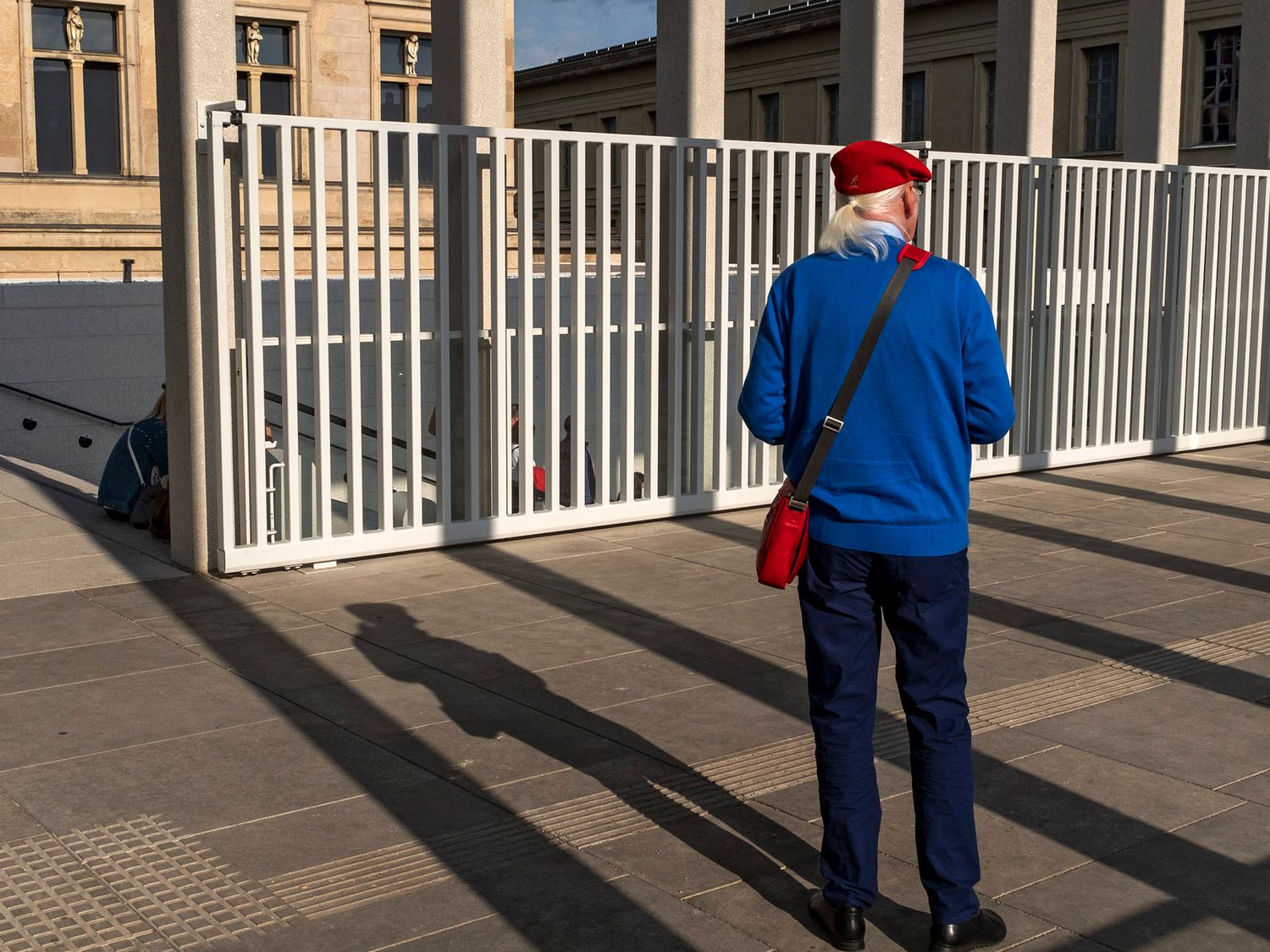 Person in blue jacket and red hat stands before white metal gates casting long shadows on pavement.