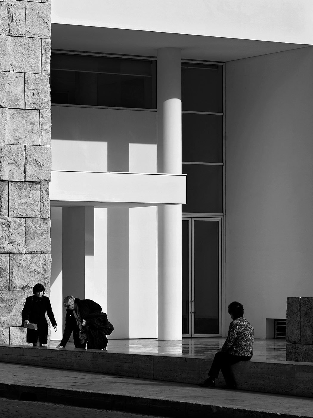 Black and white photo of people outside a modern building with geometric architecture, columns, and stone walls.