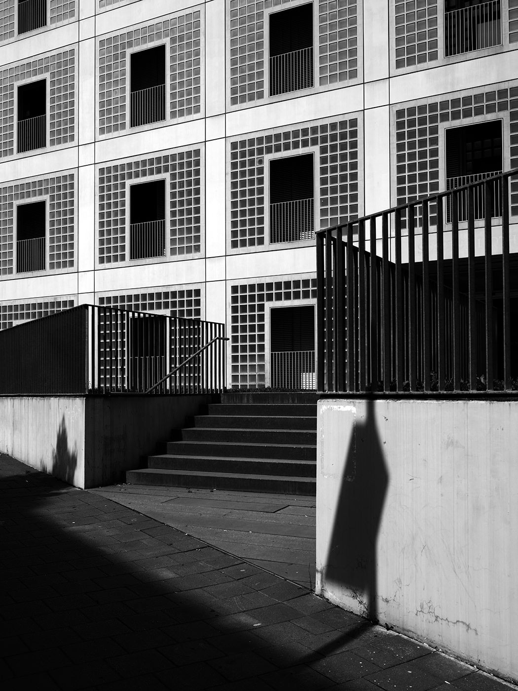 Black and white photo of a brutalist building exterior with stairs, metal railings, and dramatic shadows on concrete walls.
