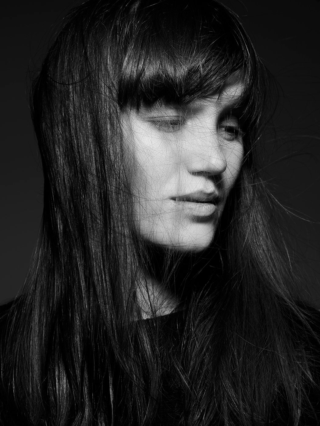 Black and white portrait of a young woman with long dark hair and bangs, eyes closed, facing down.