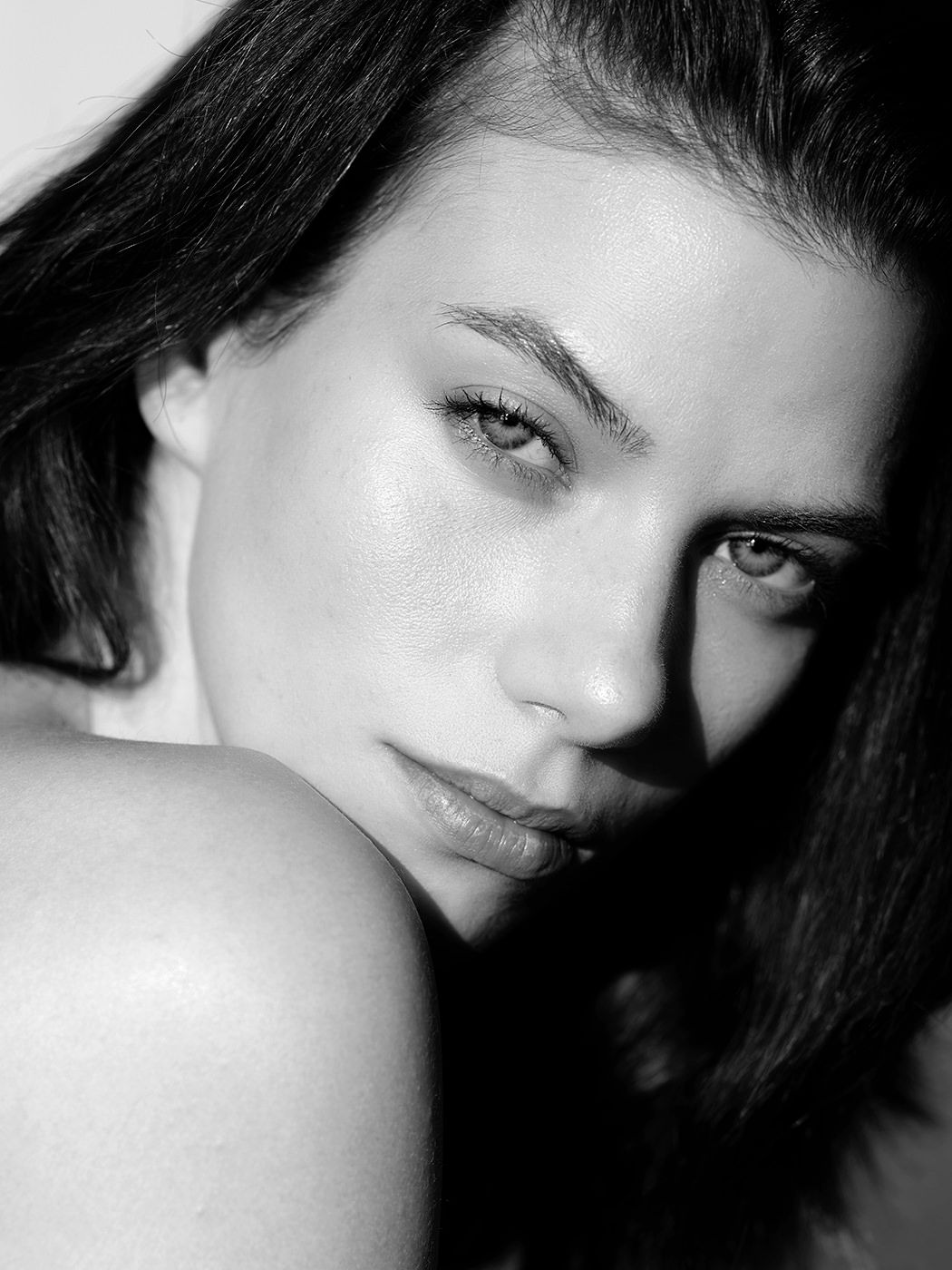 Black and white artistic portrait of a young brunette woman with bare shoulder looking intensely at camera.