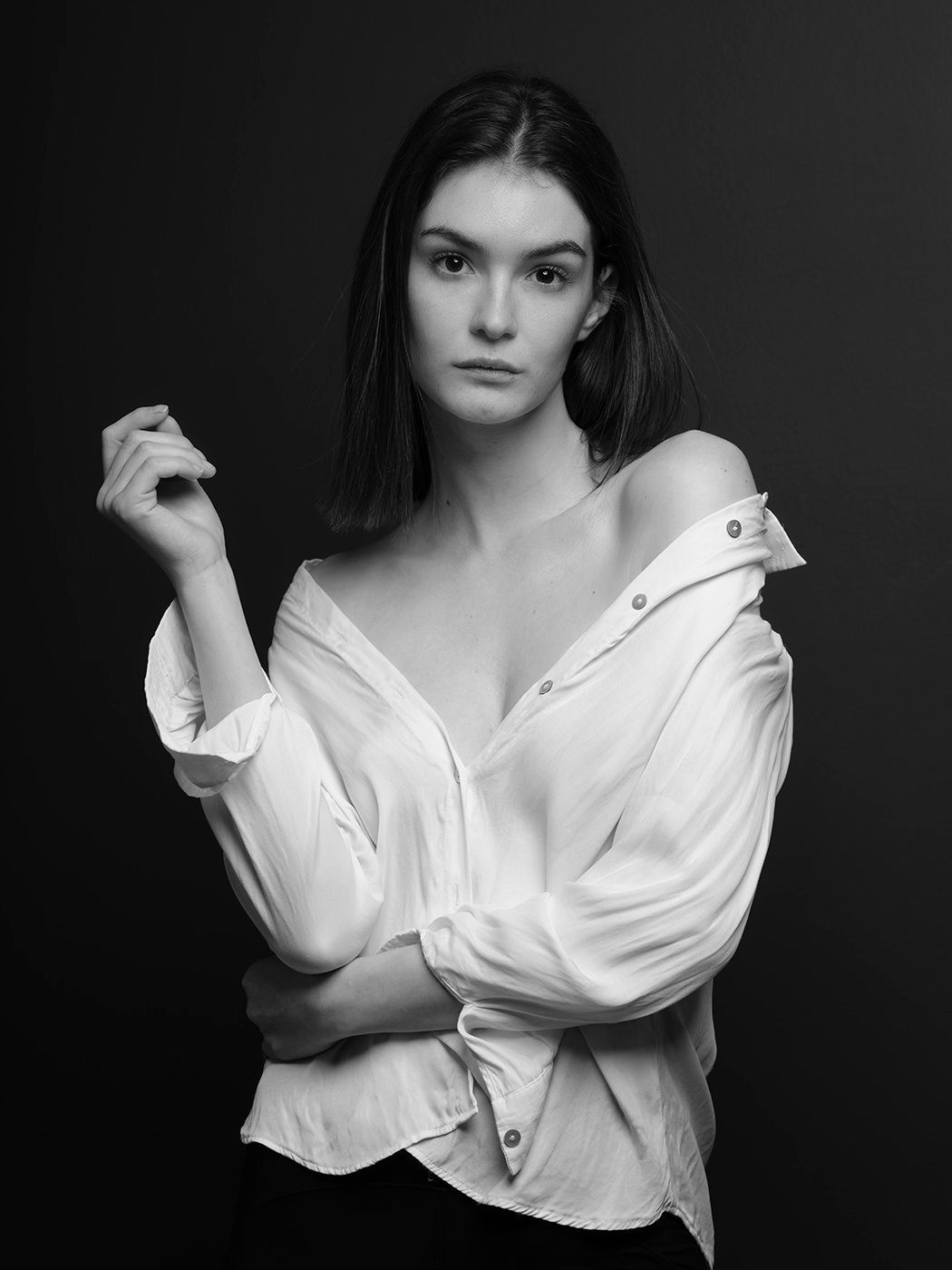 Black and white portrait of a young woman in an off-shoulder white button-up shirt posing against a dark background.