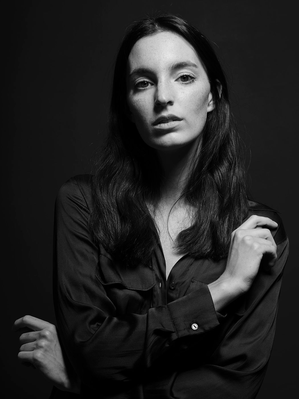 Black and white portrait of a woman with long dark hair wearing a silk shirt, arms crossed, dark background.