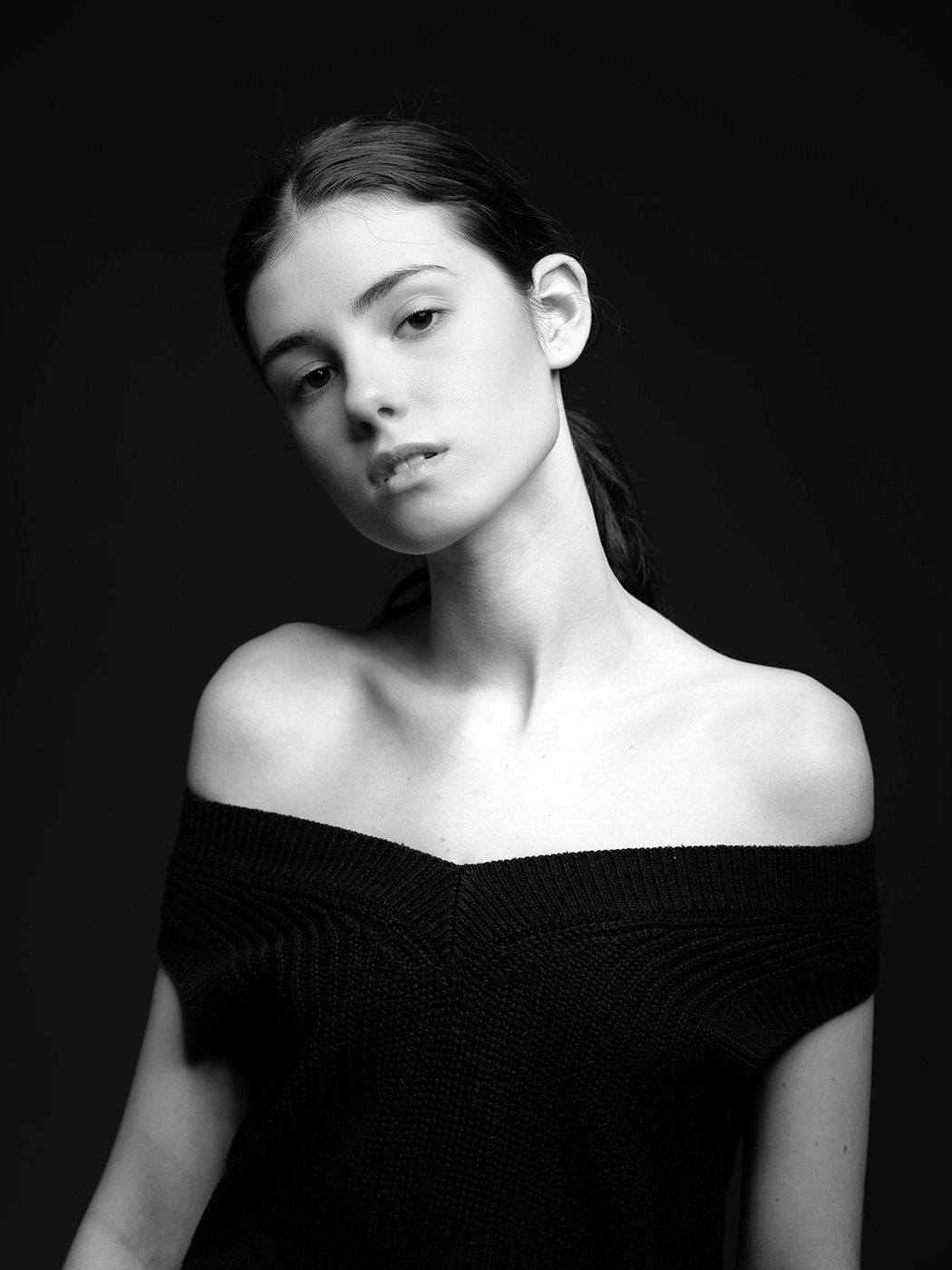 Elegant black and white portrait of a young woman in an off-shoulder black knit top against a dark background.
