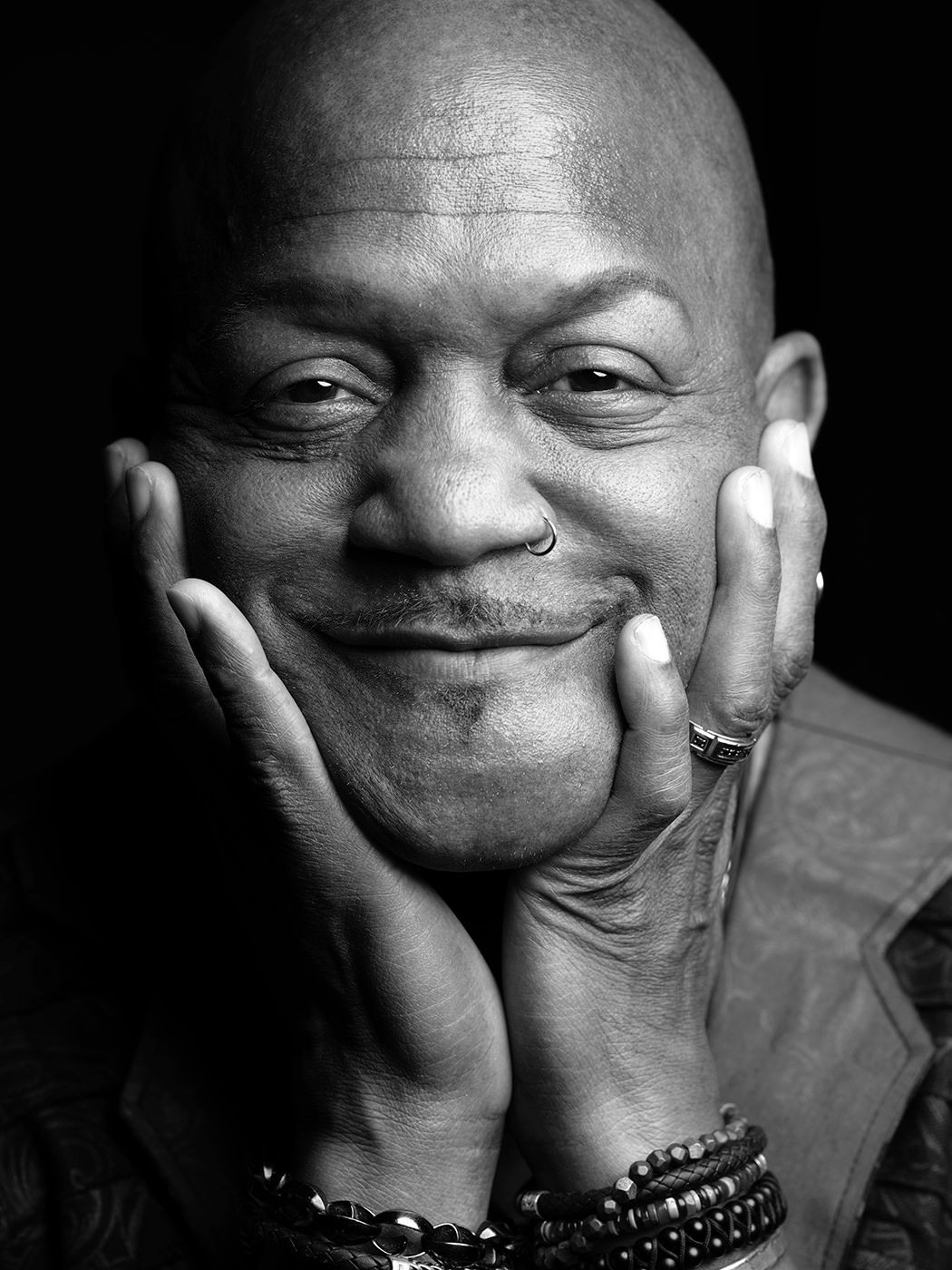 Black and white portrait of a bald man smiling, resting face in hands, wearing bracelets and a ring. Joseph Wooten