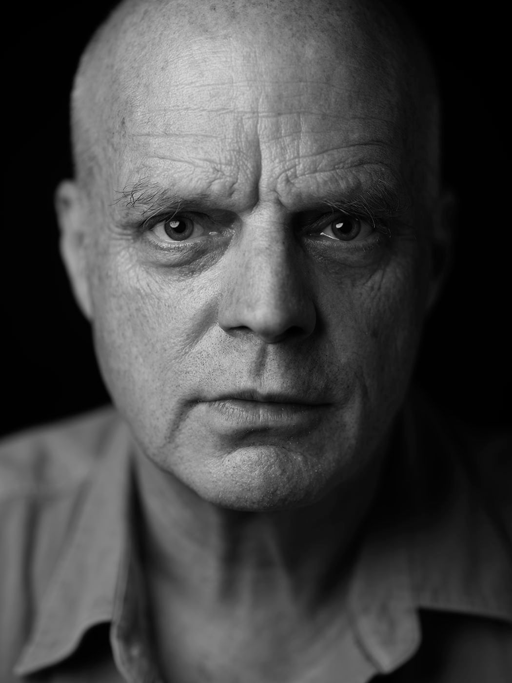 Dramatic black and white close-up portrait of a bald older man with an intense expression. John Medeski