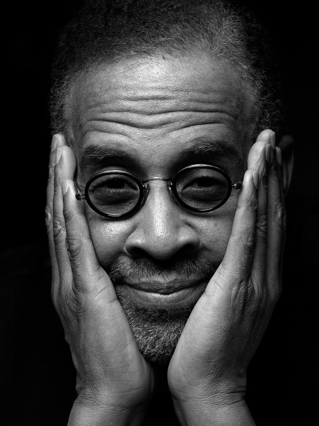 Black and white portrait of a man with round glasses, hands on cheeks, smiling warmly. Stanley Clarke, musician, bass