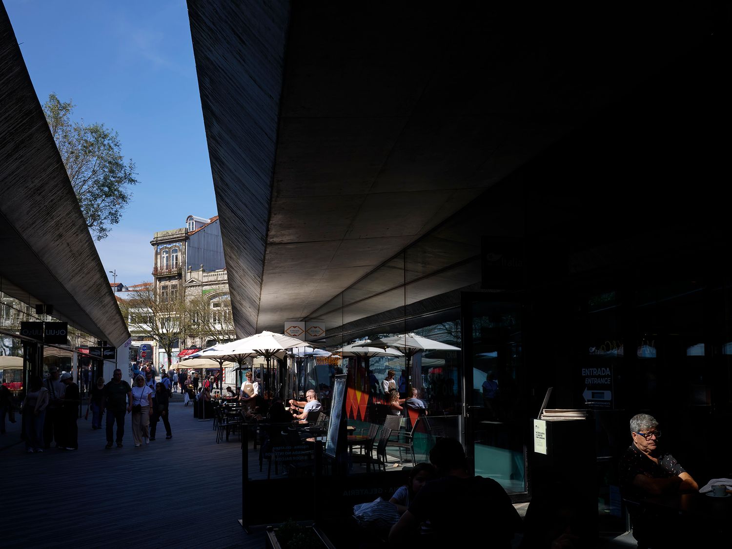 Outdoor cafe terrace under modern canopy overhang with diners and pedestrians in sunny urban plaza.