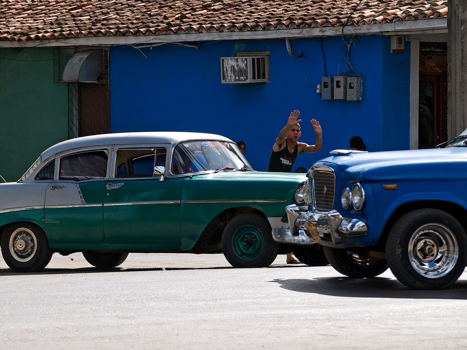 Vintage classic cars parked on a Cuban street with colorful blue building and pedestrian waving in background.