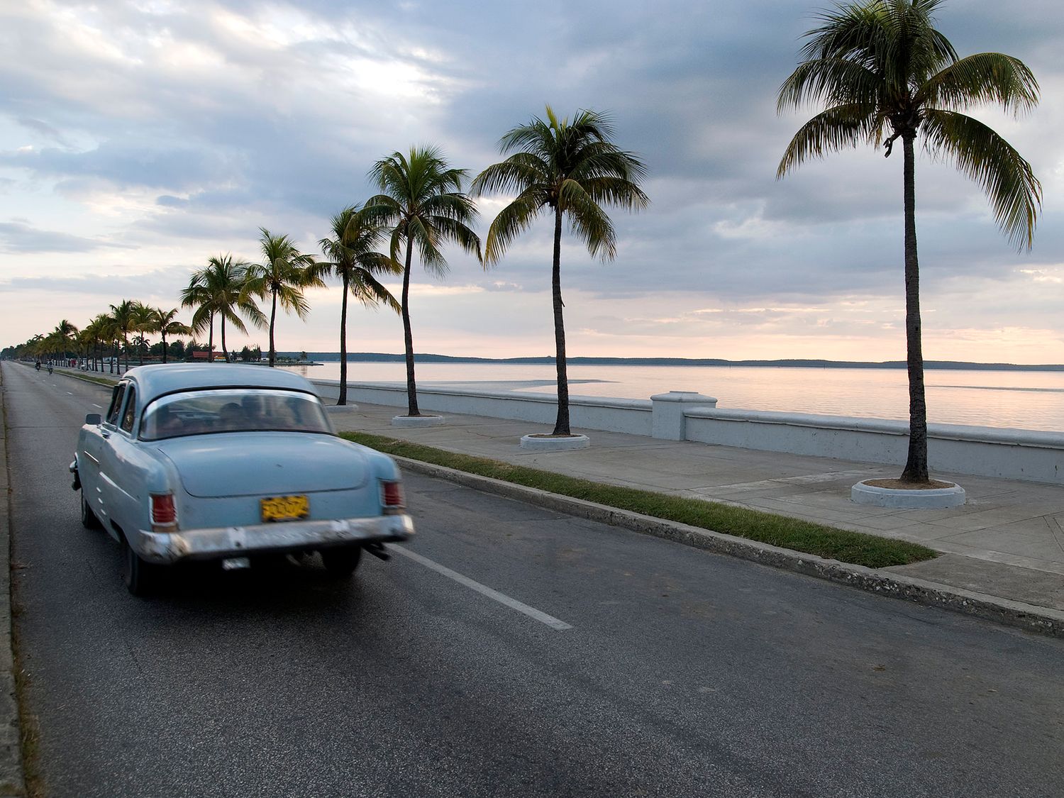 A vintage blue car drives along a palm-lined coastal road at dusk in Cuba.