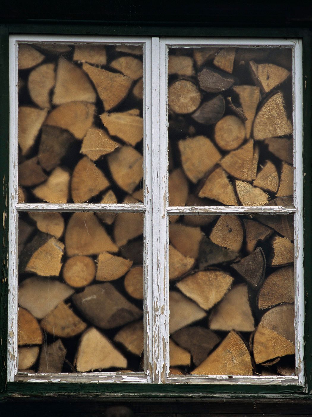 Stacked split firewood logs viewed through a rustic white-framed four-pane window of a wooden storage shed.