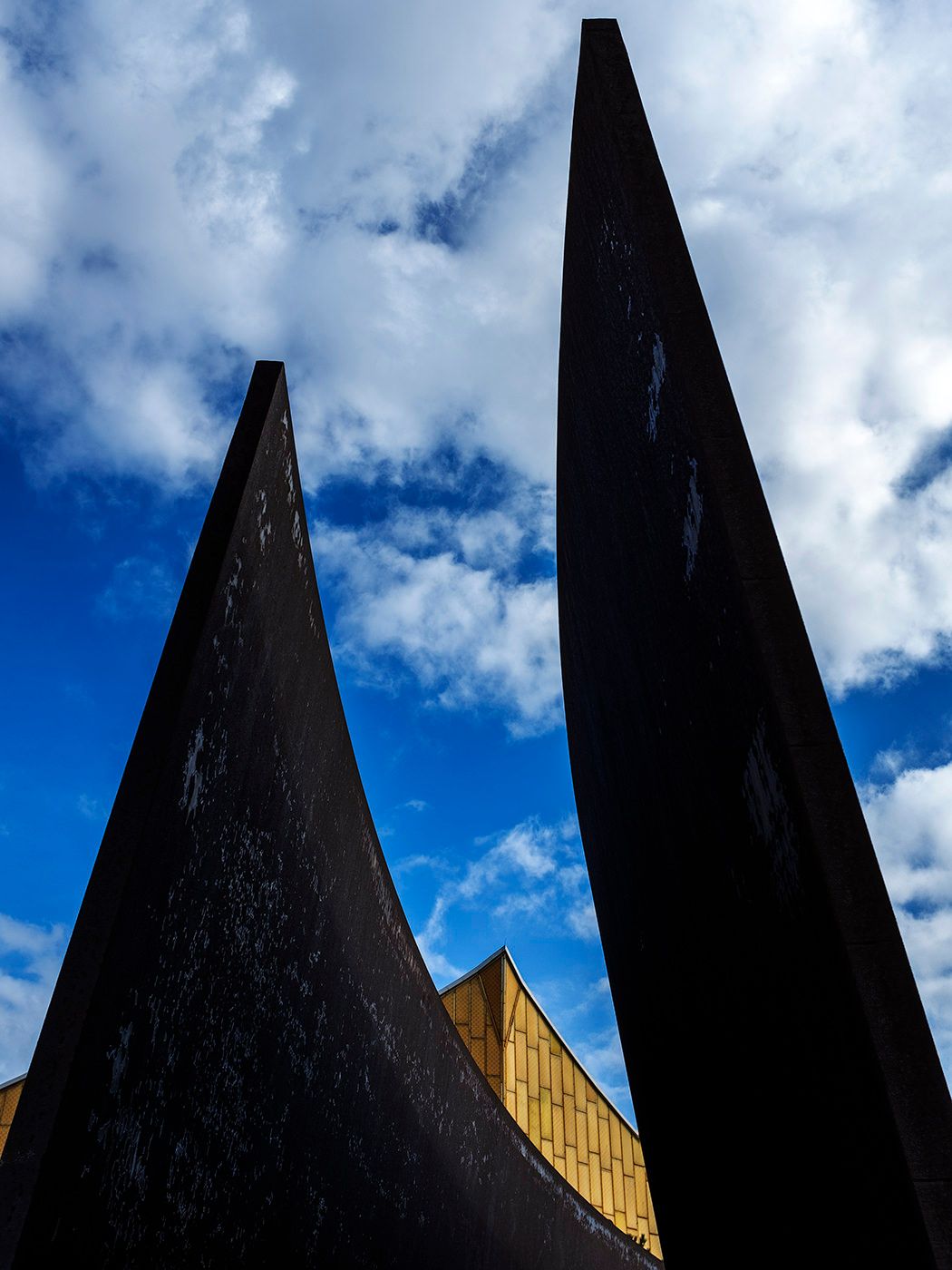Two towering dark steel sculptures rise against a vivid blue sky with white clouds and a golden building behind.