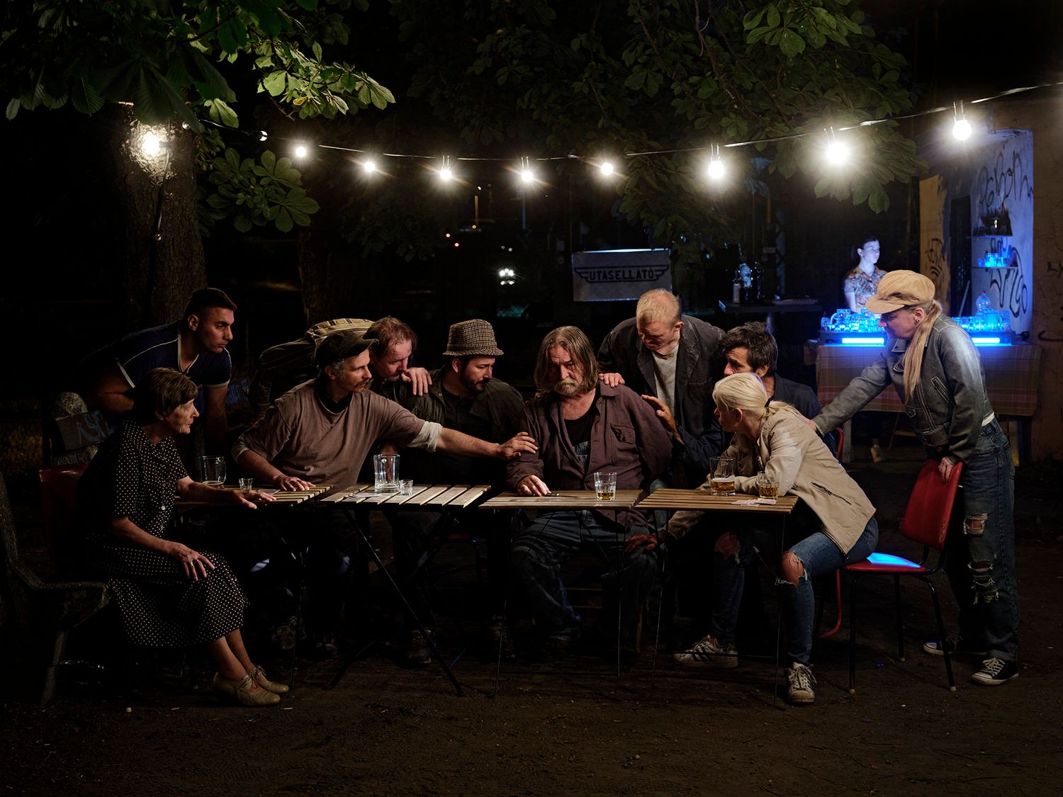 Group of people gathered around a long table outdoors at night under string lights.