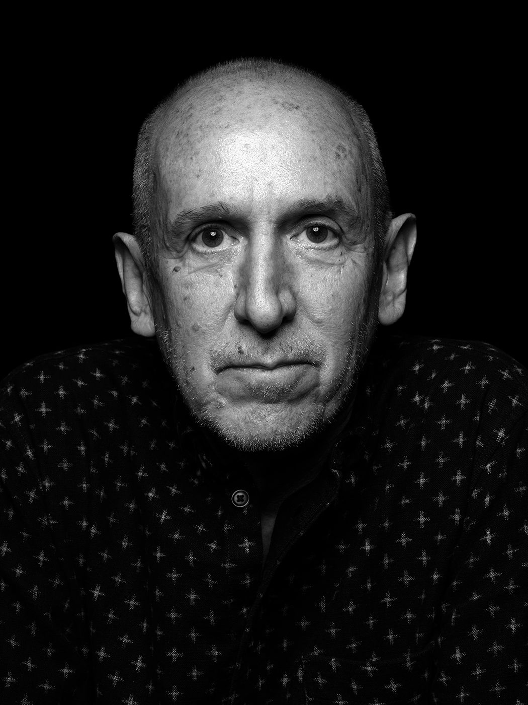 Black and white portrait of a bald man in a patterned shirt against a dark background. Michel Landau