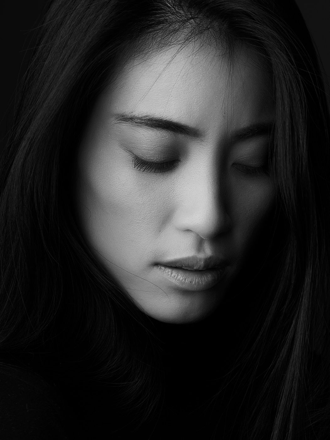 Black and white portrait of a young Asian woman with closed eyes and long dark hair against a dark background.