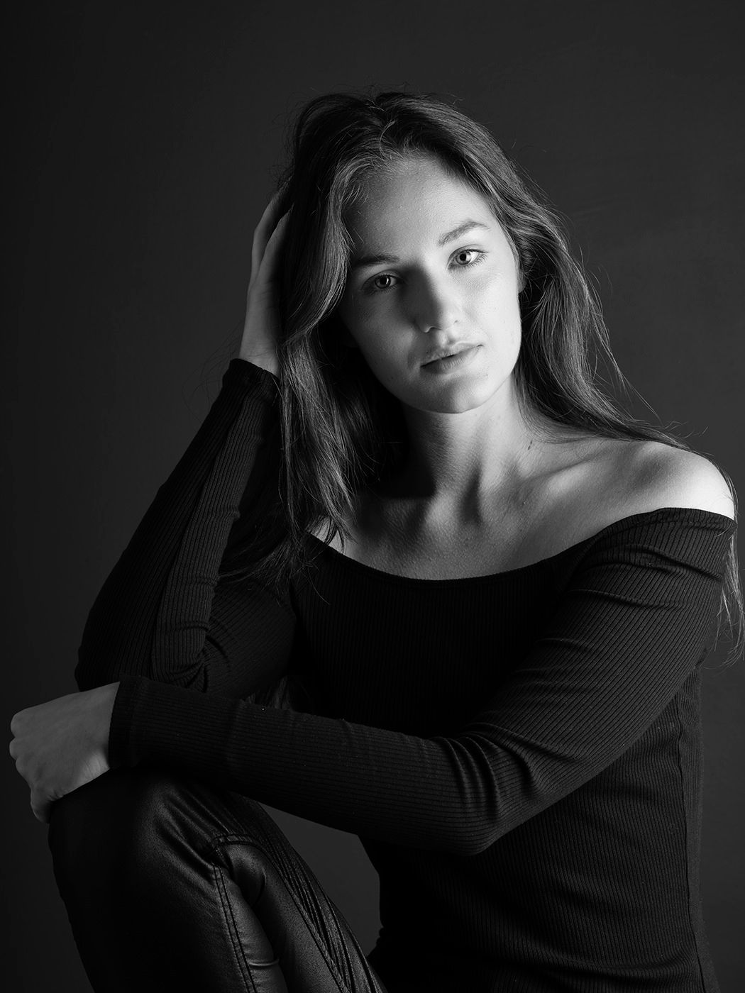 Black and white portrait of a young woman in an off-shoulder top, posing with hand in hair against dark background.
