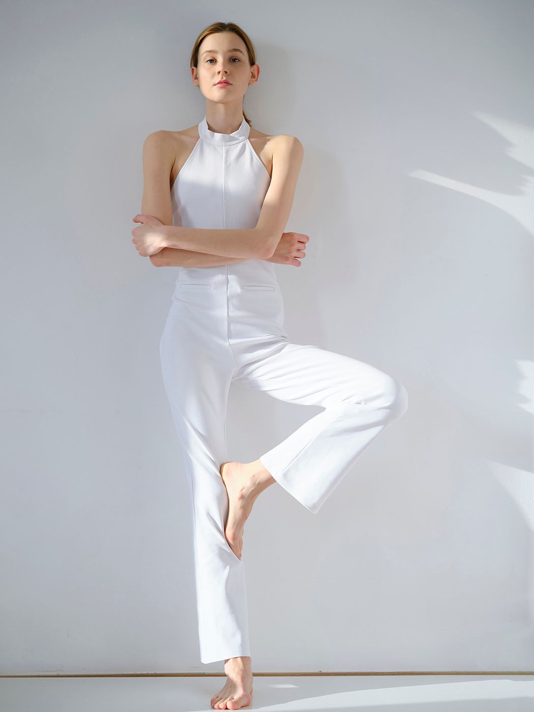 Woman in white halter jumpsuit striking a yoga tree pose against a light gray wall.