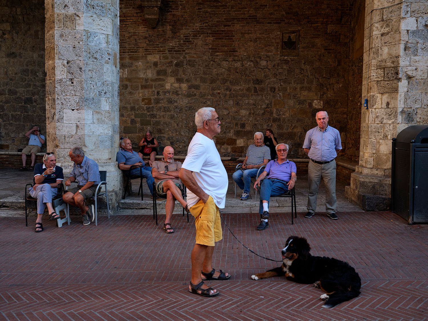 Man with dog on leash stands in Italian piazza while locals relax in chairs under stone archways.