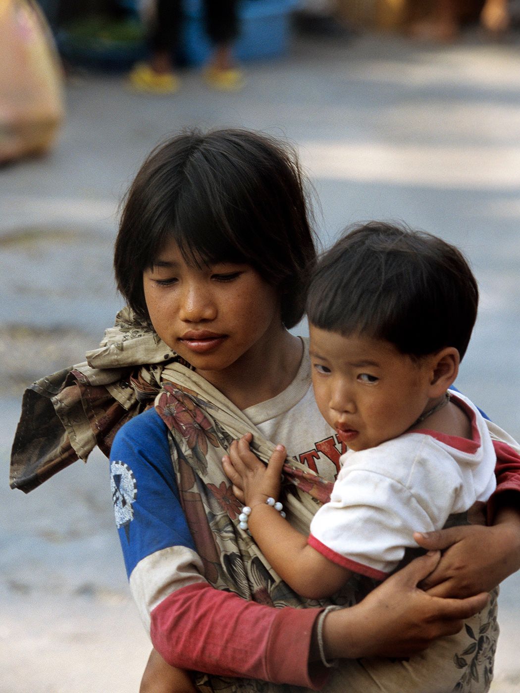 Young Asian girl carrying a toddler on her back using a traditional cloth wrap on a street.