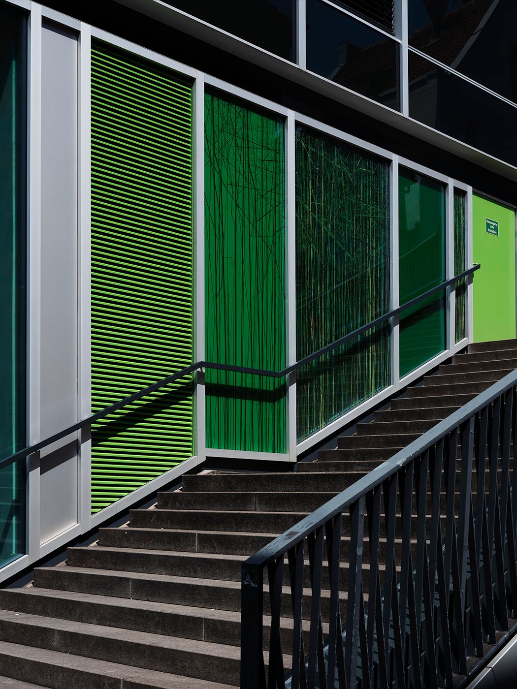 Modern building exterior with green panels and black railings on concrete stairs in bright sunlight.