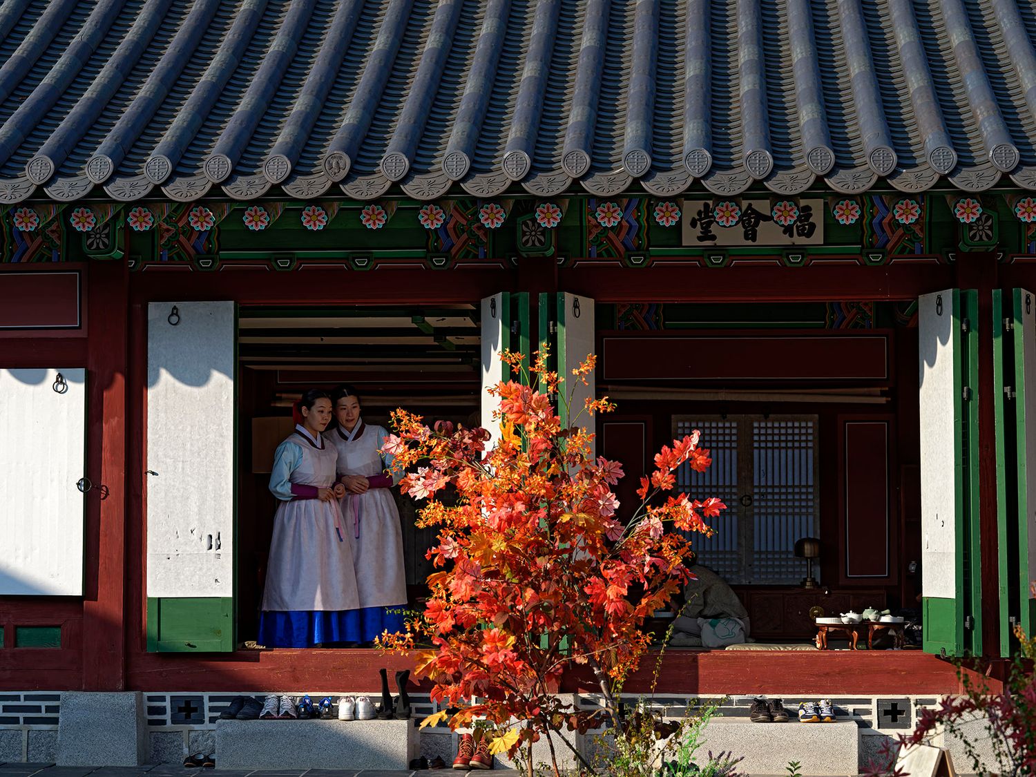 Two women in traditional Korean hanbok stand in a colorful temple doorway framed by vibrant autumn foliage.