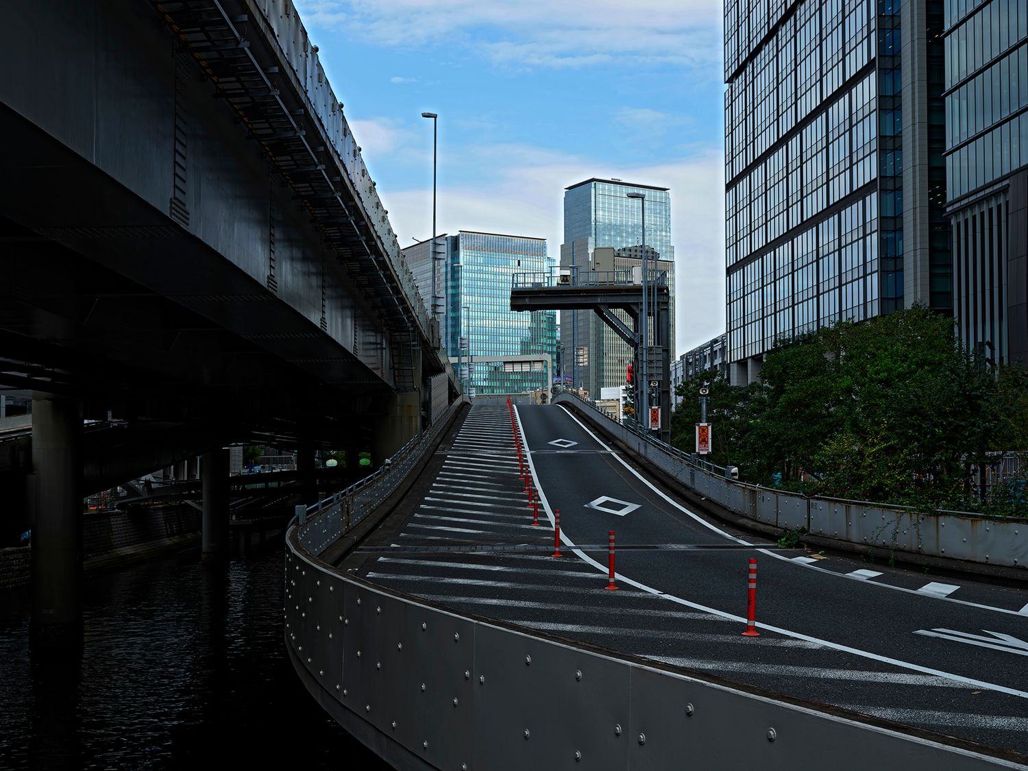 Curved urban highway with red lane markings beside a waterway, elevated expressway overhead, modern glass buildings in Tokyo.