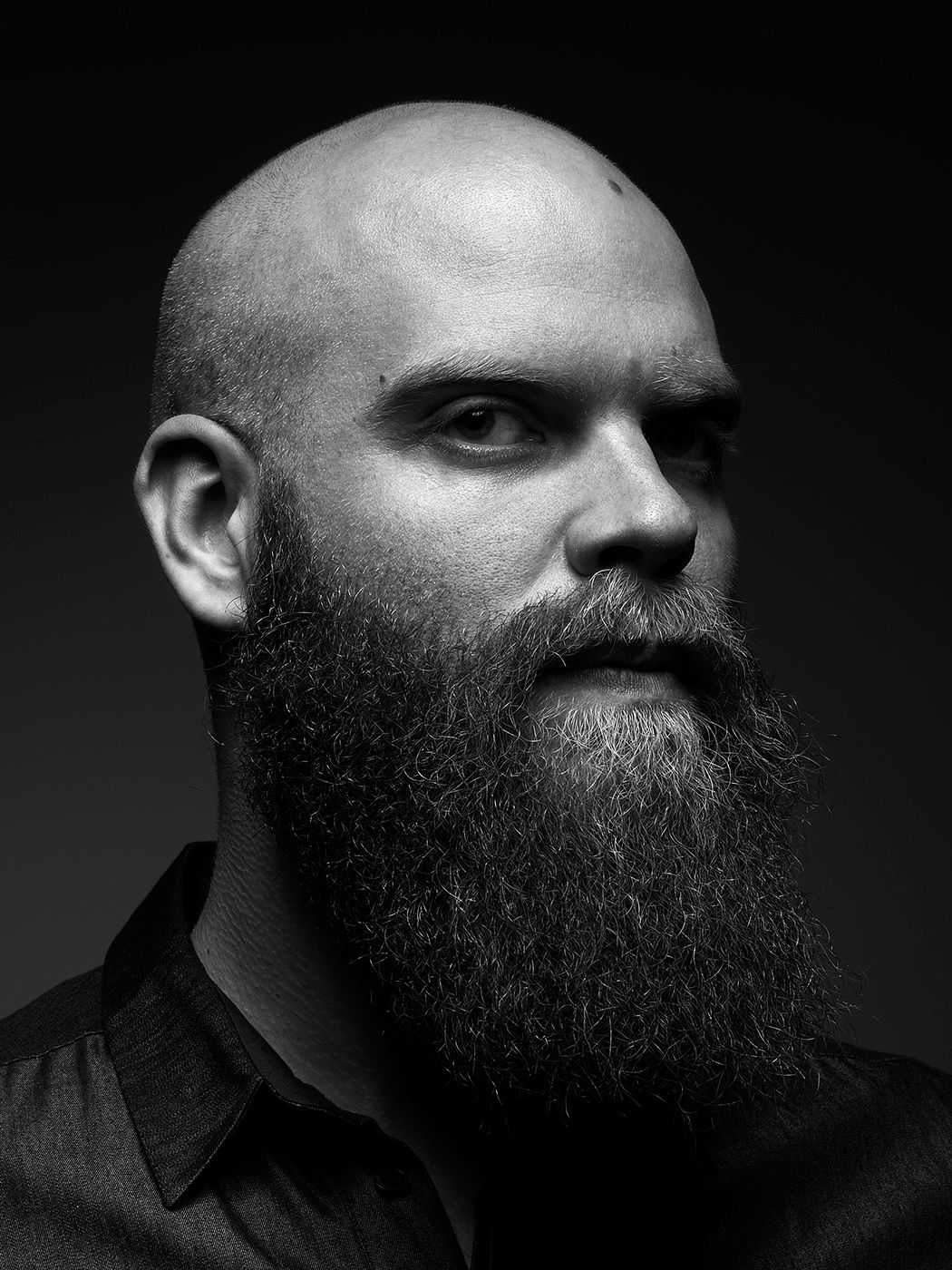 Black and white portrait of a bald man with a long, full beard wearing a dark shirt against a dark background. Jobbágy Bence