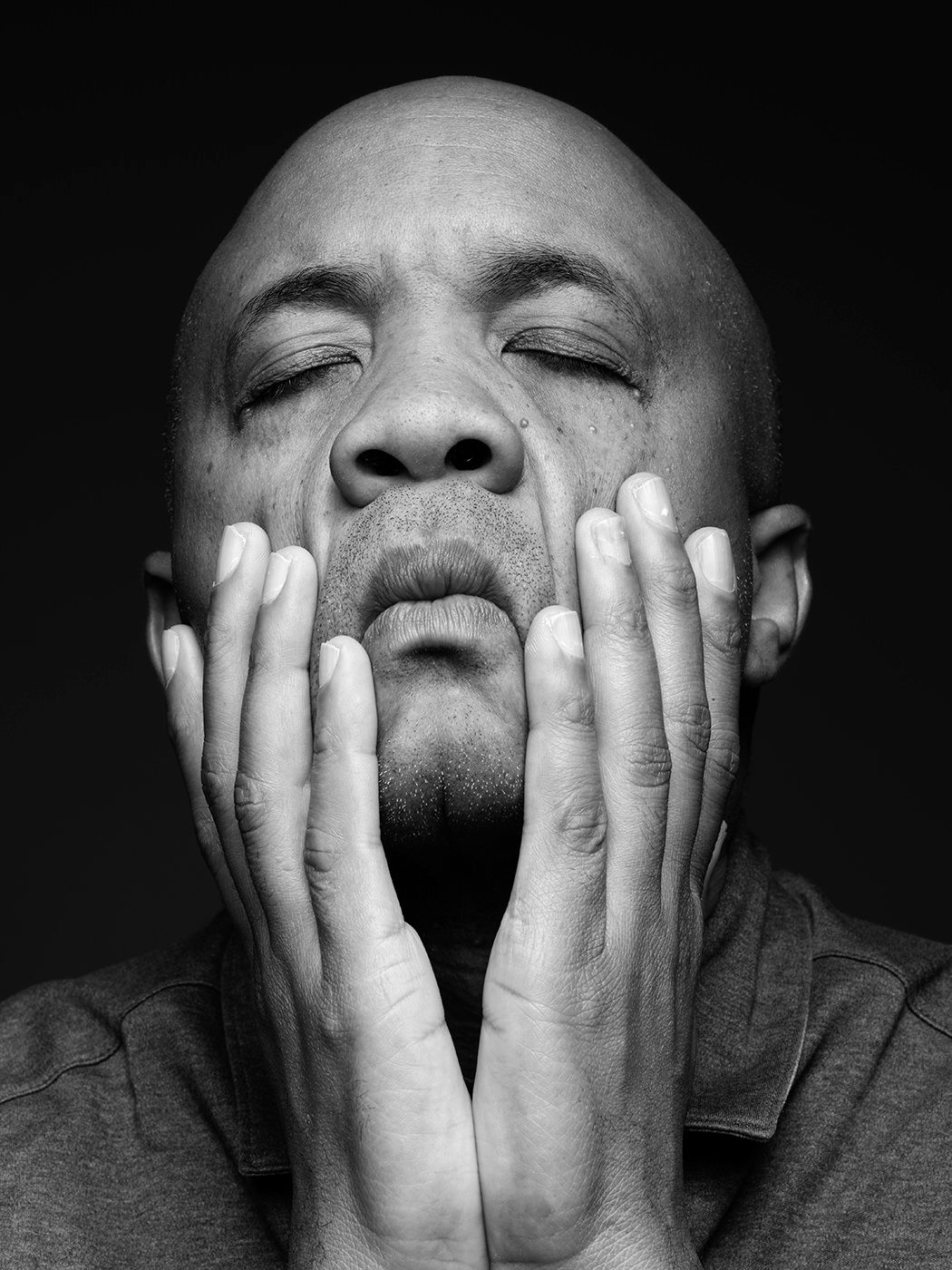 Black and white portrait of a bald man with eyes closed, hands cupping his face in a contemplative pose. Reuben Rogers, musician, bass.