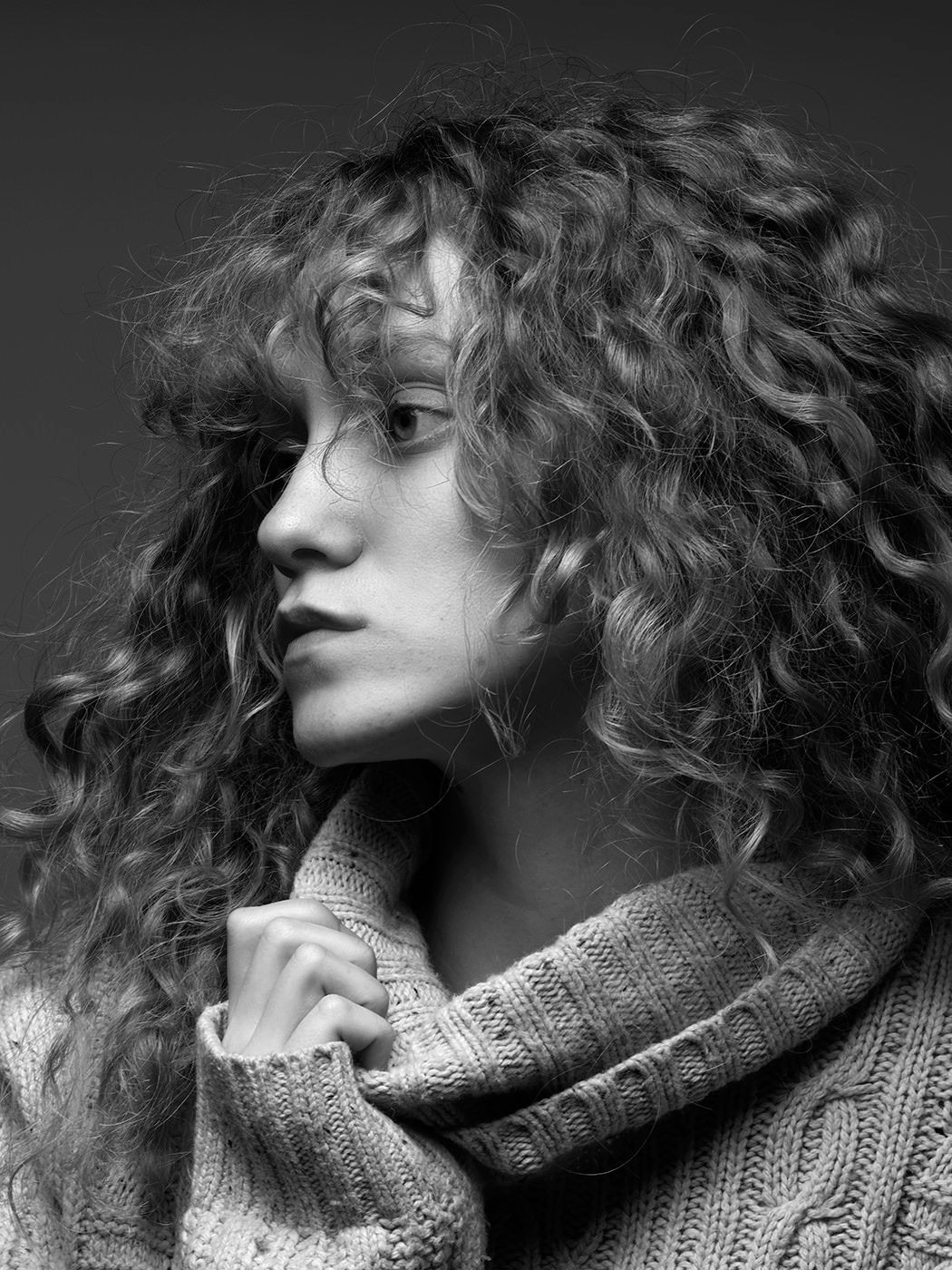Black and white portrait of a young woman with voluminous curly hair wearing a cozy knit turtleneck sweater.
