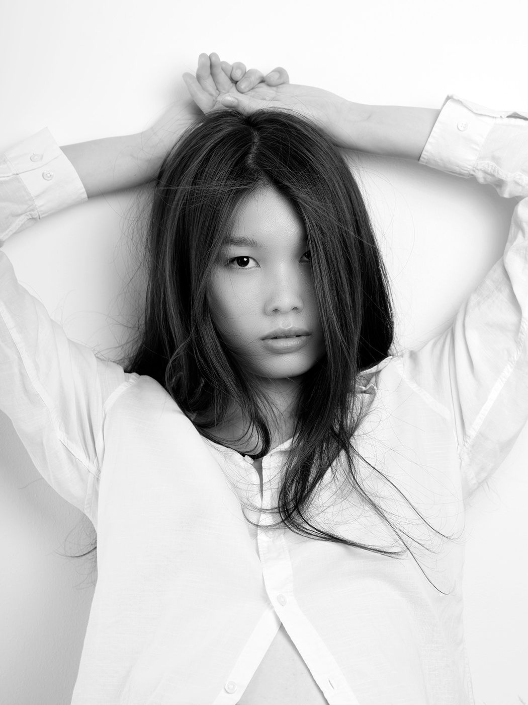 Black and white portrait of a young Asian woman in a white shirt, arms raised behind her head, gazing at camera.