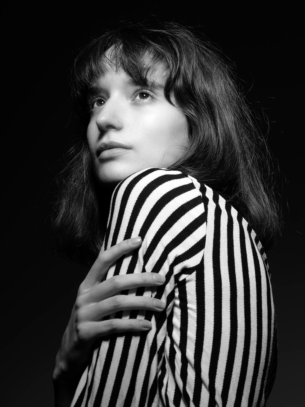 Black and white portrait of a young woman in a striped top, gazing over her shoulder against a dark background.