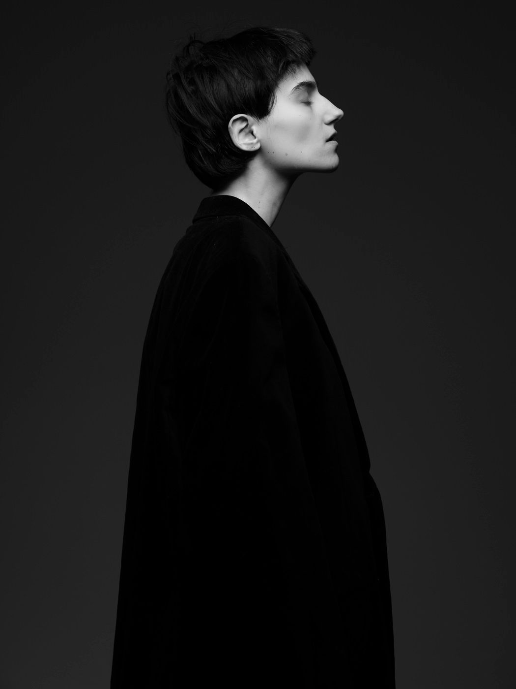 Black and white profile portrait of a young person in an oversized dark coat, eyes closed, against a dark background.
