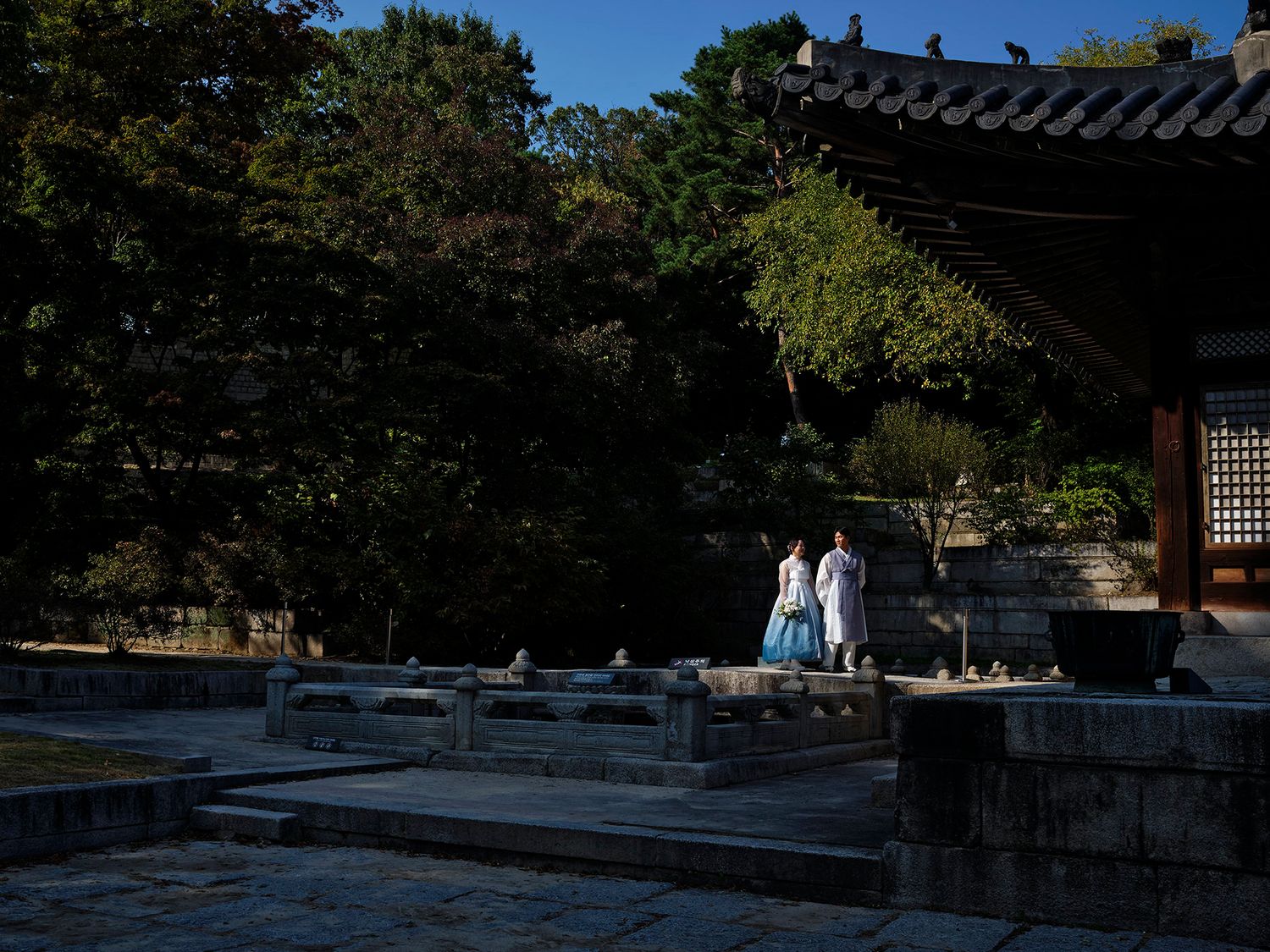 Two people in traditional Korean hanbok attire stand at a historic palace courtyard surrounded by lush trees.