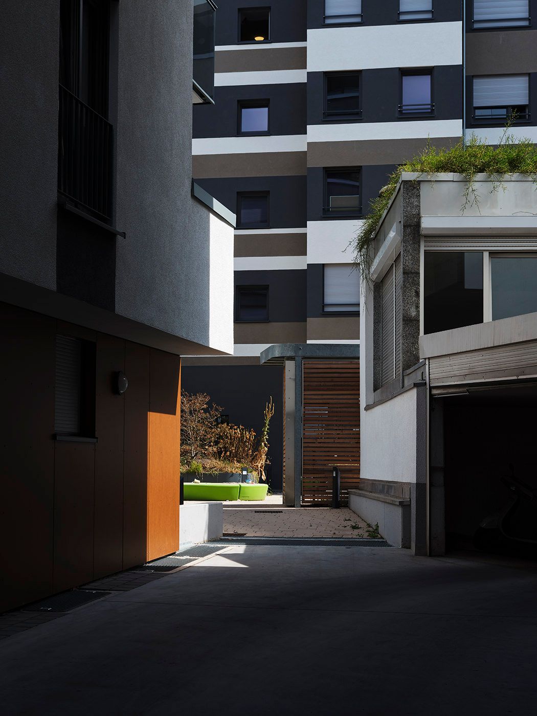 Narrow urban courtyard between modern apartment buildings with orange wall accent and green seating area in sunlight.