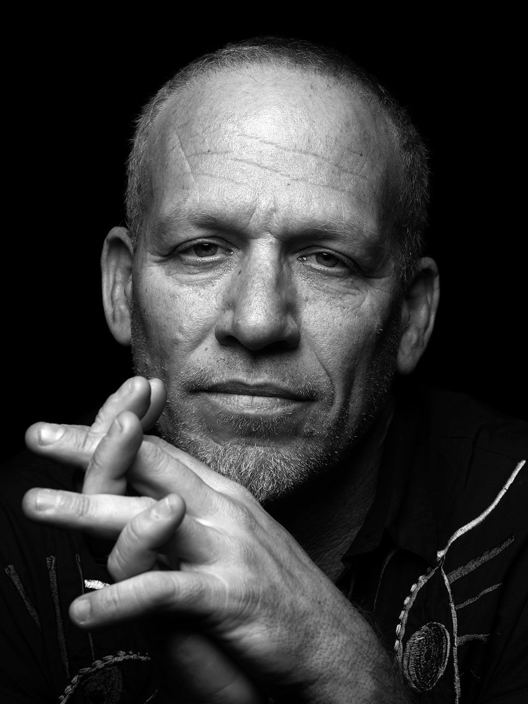 Black and white portrait of a bald man with hands clasped under chin, intense gaze, dramatic lighting. Avishai Cohen