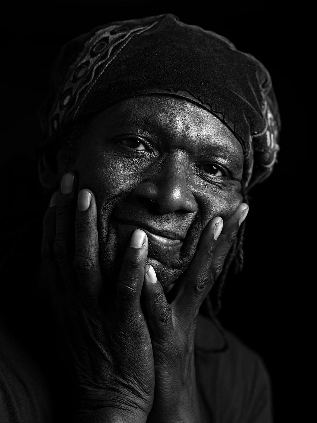 Black and white portrait of a smiling elder with hands on cheeks, wearing a patterned headwrap. Hamid Drake