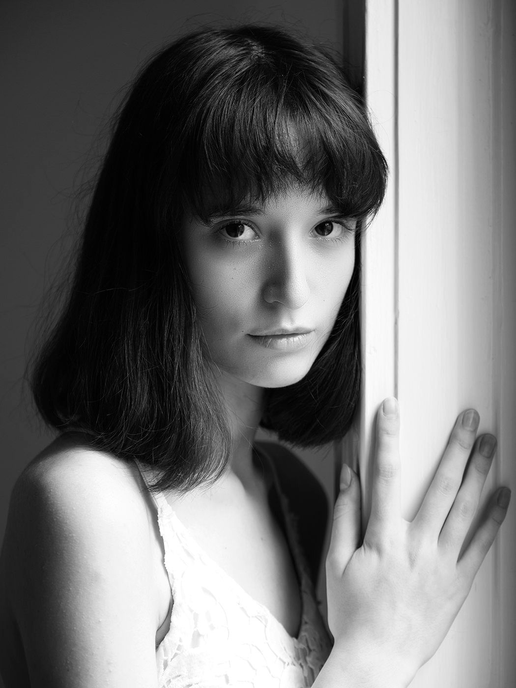 Black and white portrait of a young woman with dark hair and bangs leaning against a window frame.