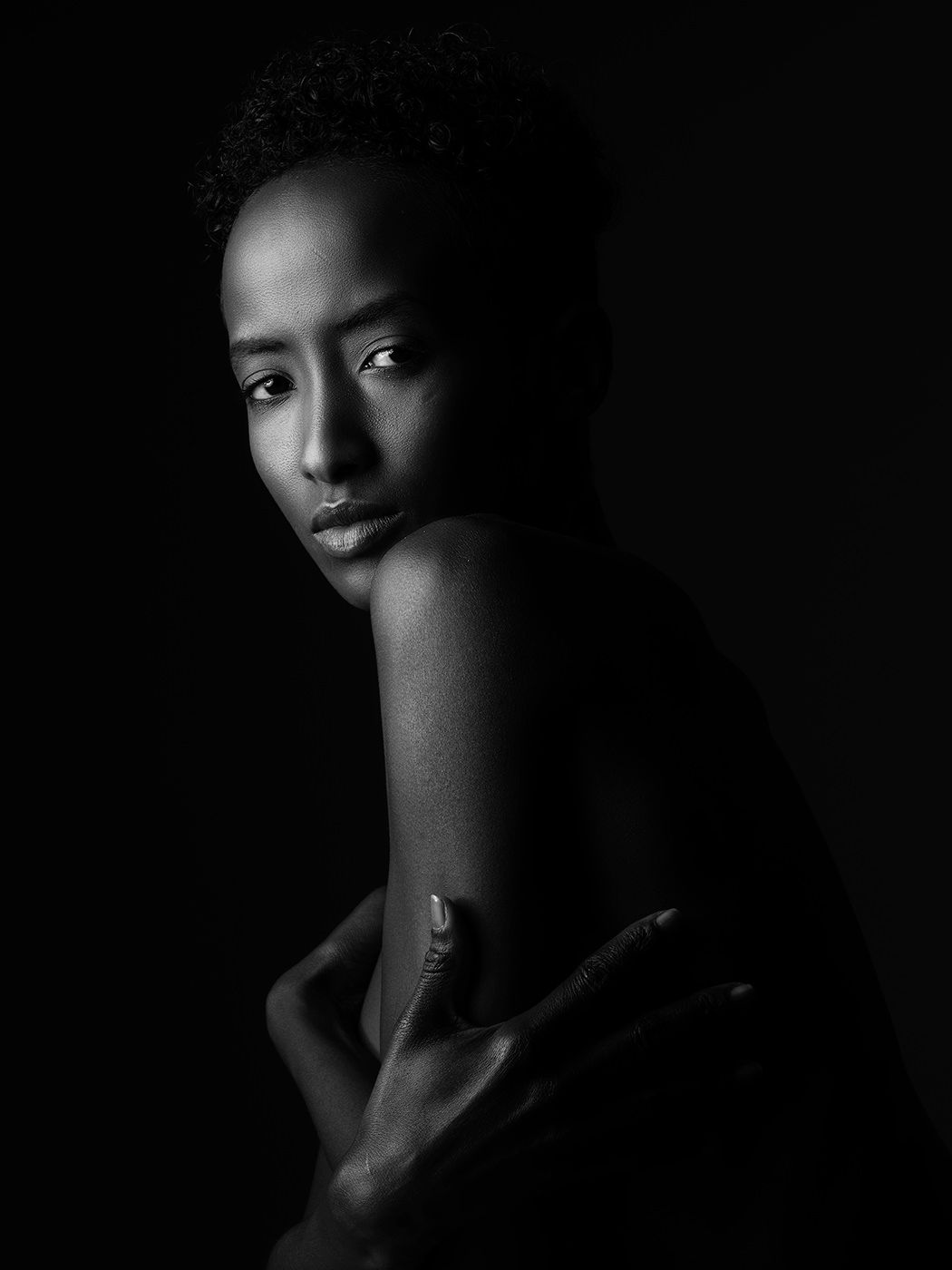 Black and white portrait of a young woman with short hair looking over her shoulder against a dark background.