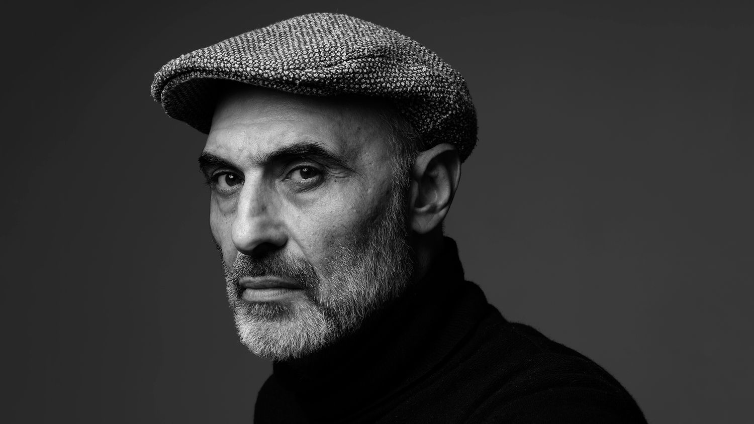 Black and white portrait of a distinguished older man wearing a flat cap and black turtleneck.