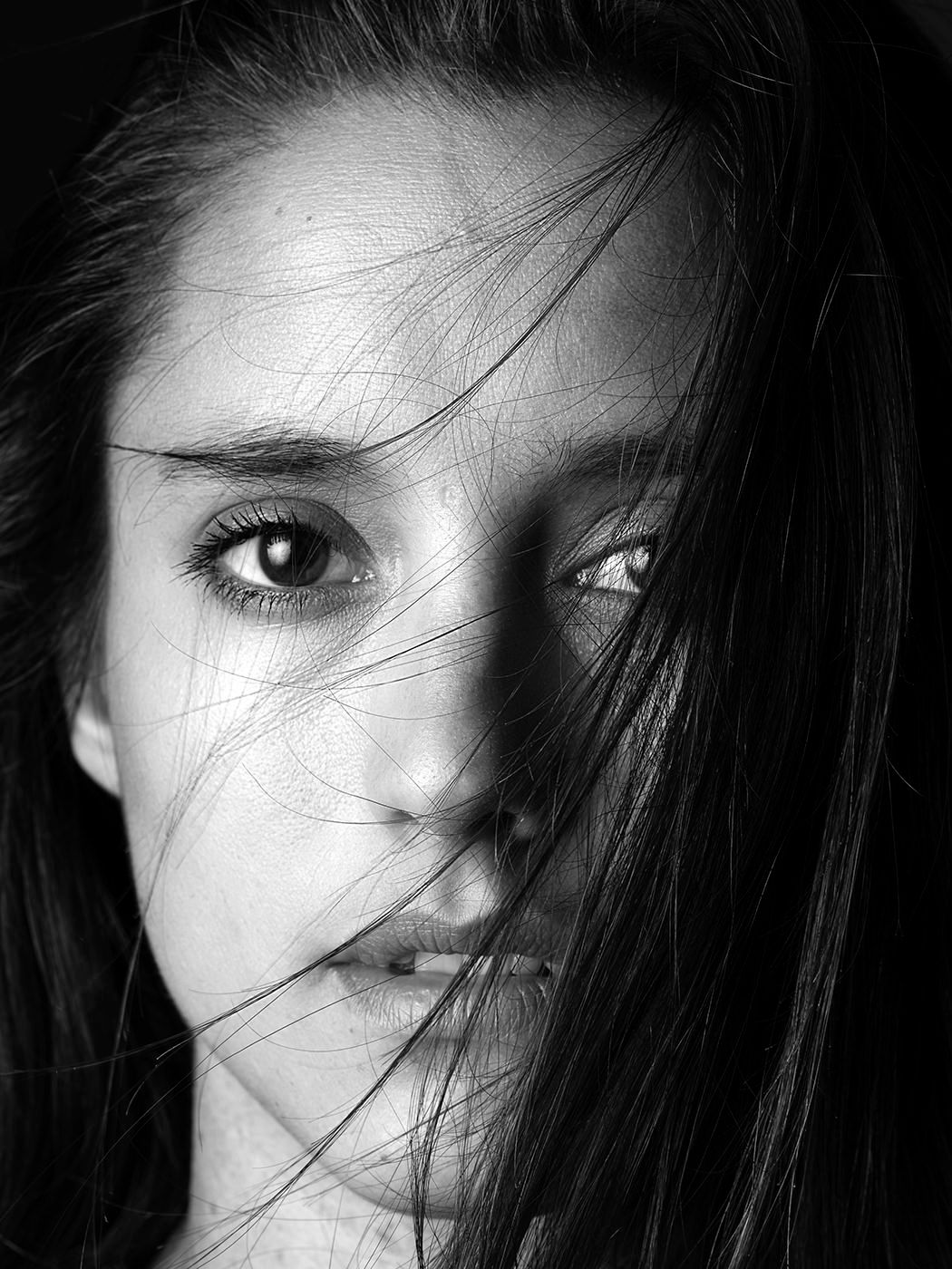 Dramatic black and white close-up portrait of a young woman with dark hair blowing across her face.