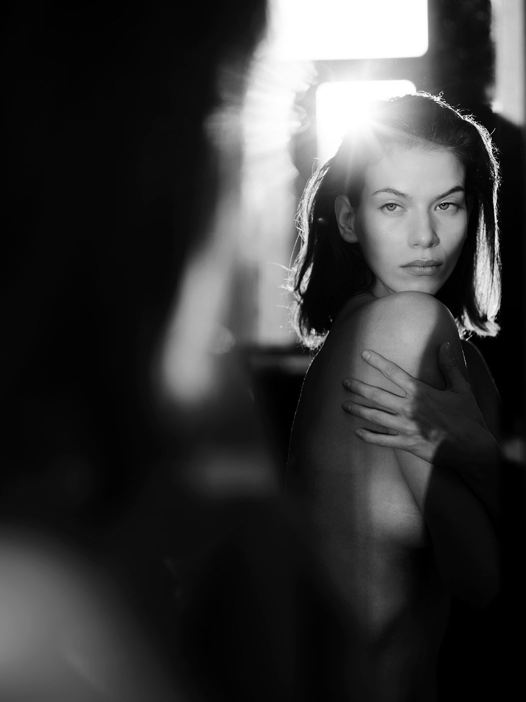 Black and white portrait of a woman with backlit glow, arms crossed, gazing forward.