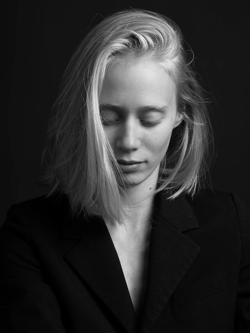 Black and white portrait of a blonde woman in a dark blazer, eyes closed, against a dark background.