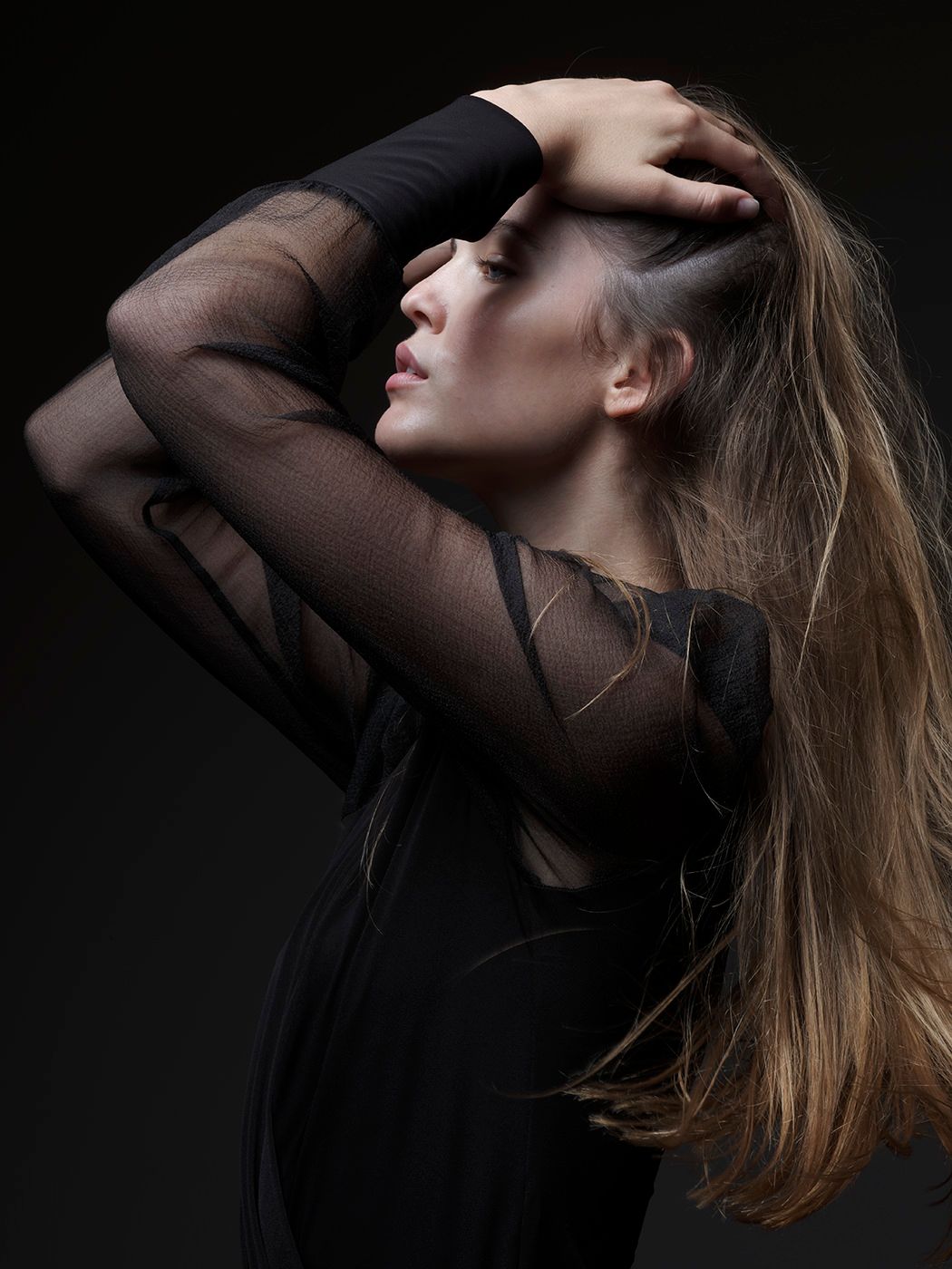 Woman in black sheer top poses dramatically with hands on head, long hair flowing, against dark background.