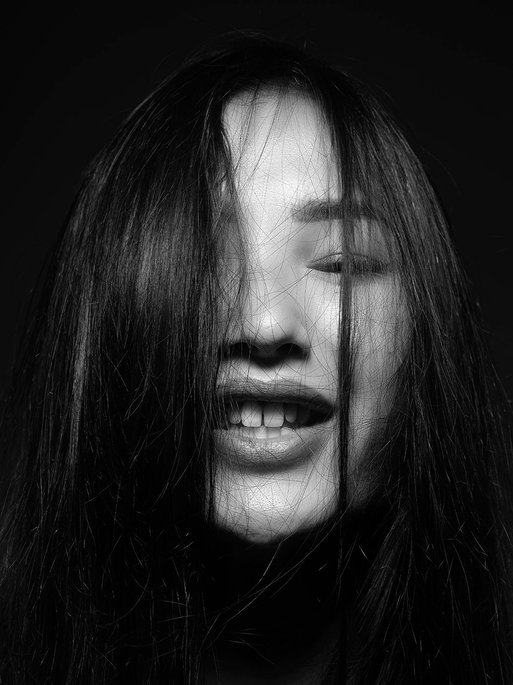 Black and white portrait of a woman with long hair covering her face, mouth slightly open, dramatic dark background.