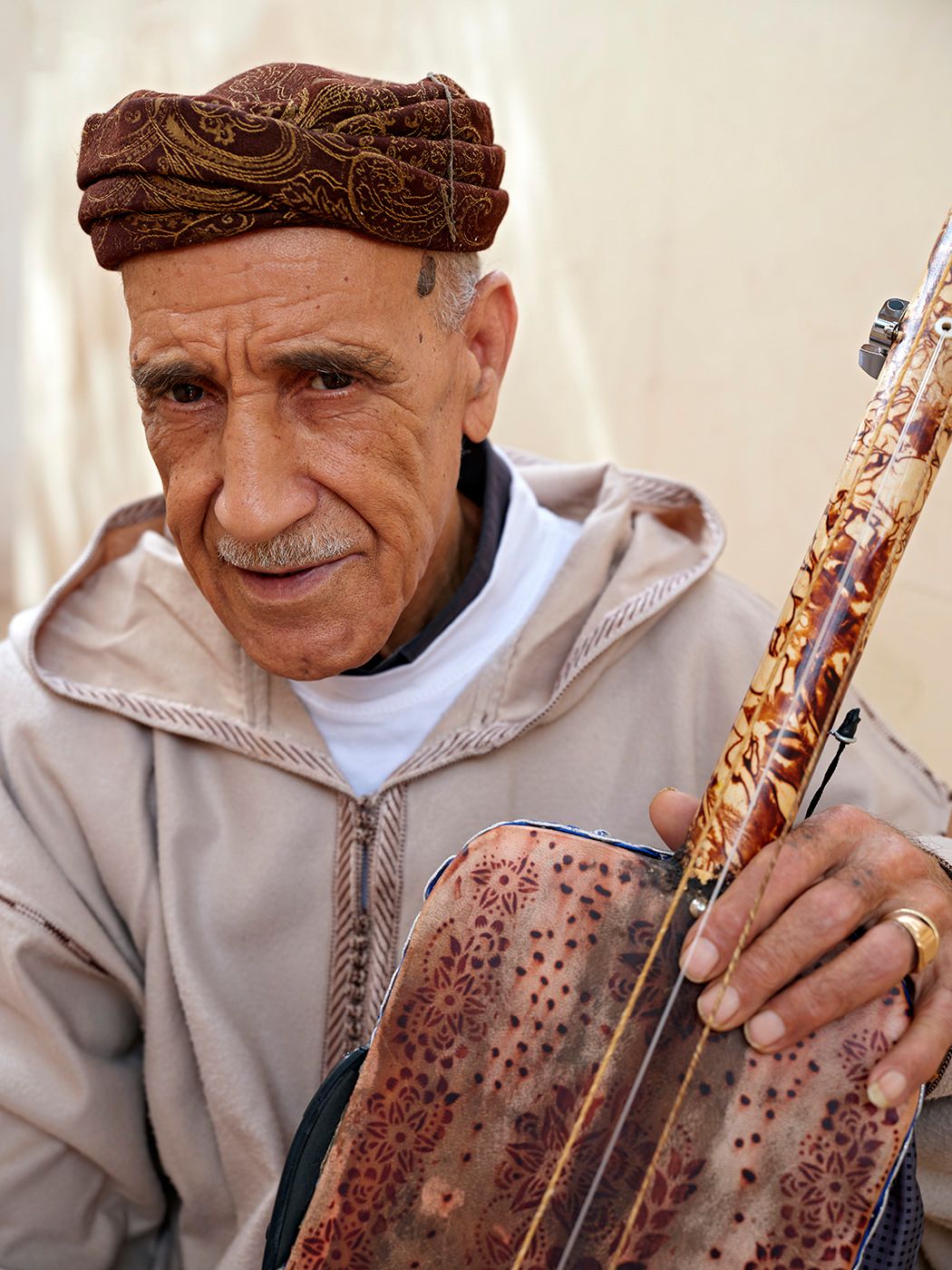 Elderly Moroccan man in traditional attire holding an ornate stringed instrument, wearing a decorative red turban.
