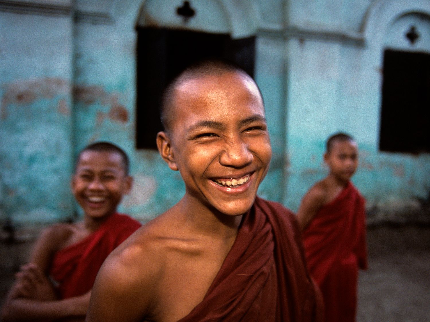 Smiling young Buddhist monks in red robes stand together outside a temple in Myanmar.