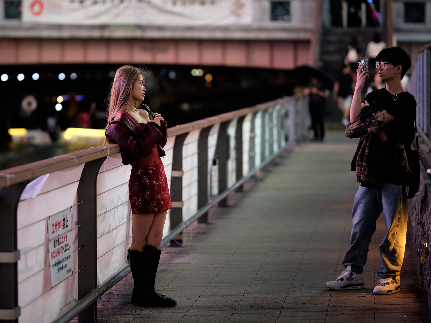 Young woman in red dress poses for photo on urban bridge at night while man photographs her.